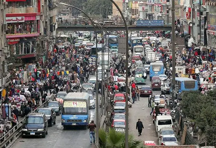 Rua movimentada no Cairo com edifícios de tijolos aparentes e pessoas caminhando, representando o cotidiano de comunidades migrantes.