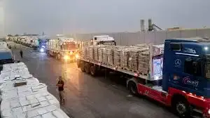 Fileira de caminhões brancos com bandeiras do Egito e emblemas humanitários cruzando um portão de segurança em zona desértica.