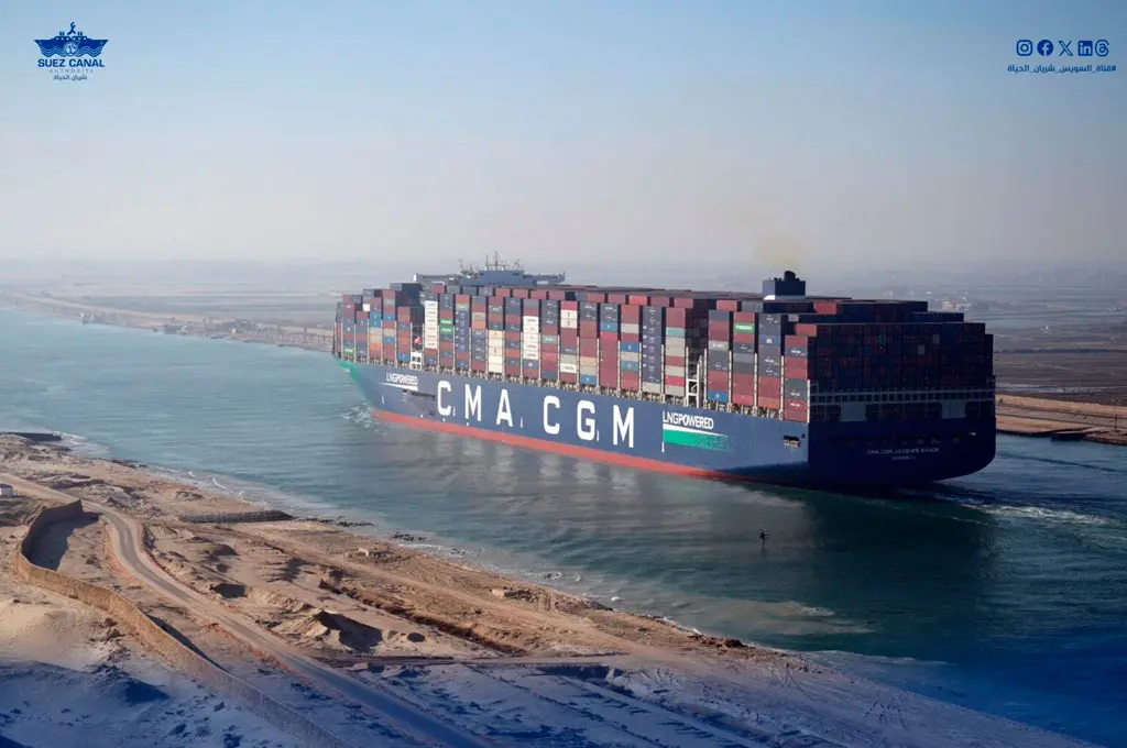 Fileira de grandes navios de carga navegando pelo Canal de Suez sob uma luz alaranjada de fim de tarde.